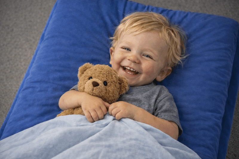 Lachendes Kind mit Teddy auf der Schlafmatratze